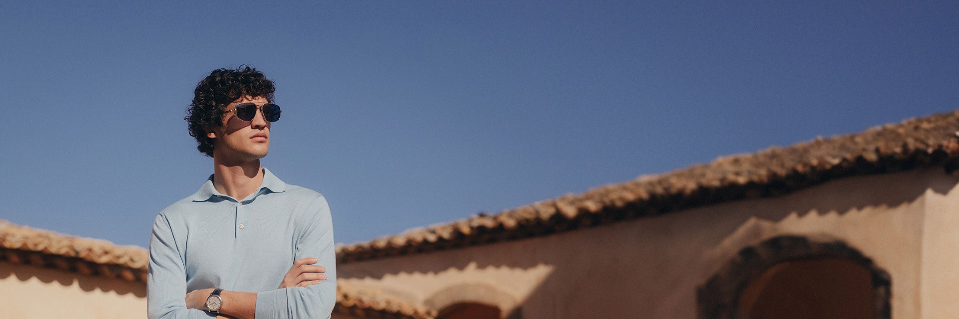 Mann mit hellblauem Langarm-Poloshirt, gold-dunkelblauer Aviator-Sonnenbrille vor Haus mit Ziegeldach und blauem Himmel. Mann mit hellblauem Langarm-Poloshirt, gold-dunkelblauer Aviator-Sonnenbrille vor Haus mit Ziegeldach und blauem Himmel.