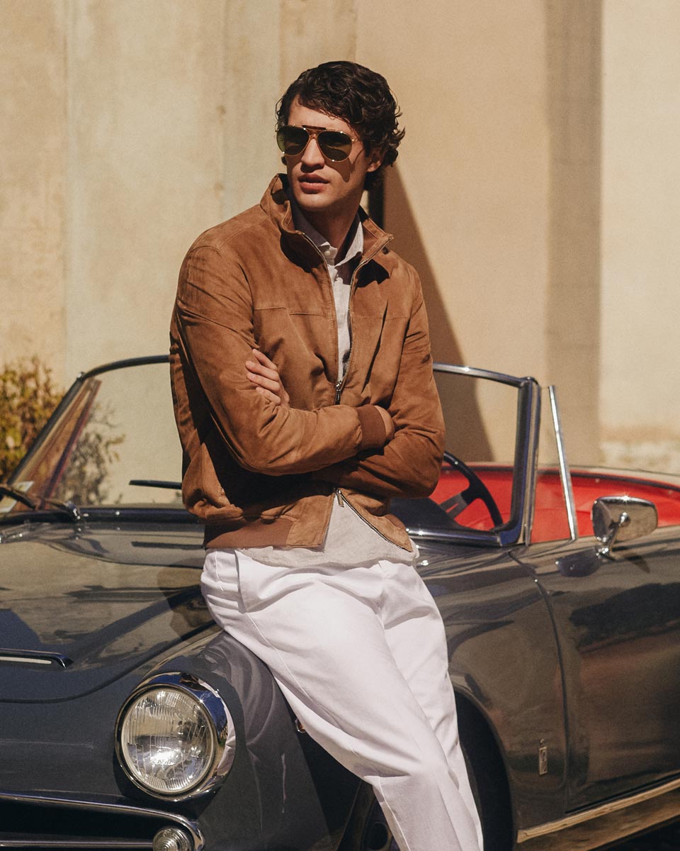 Mann mit brauner Velourslederjacke, beigem Strickpolo, weißer Hose und Sonnenbrille lehnt an grauem Oldtimer Cabrio. Mann mit brauner Velourslederjacke, beigem Strickpolo, weißer Hose und Sonnenbrille lehnt an grauem Oldtimer Cabrio.