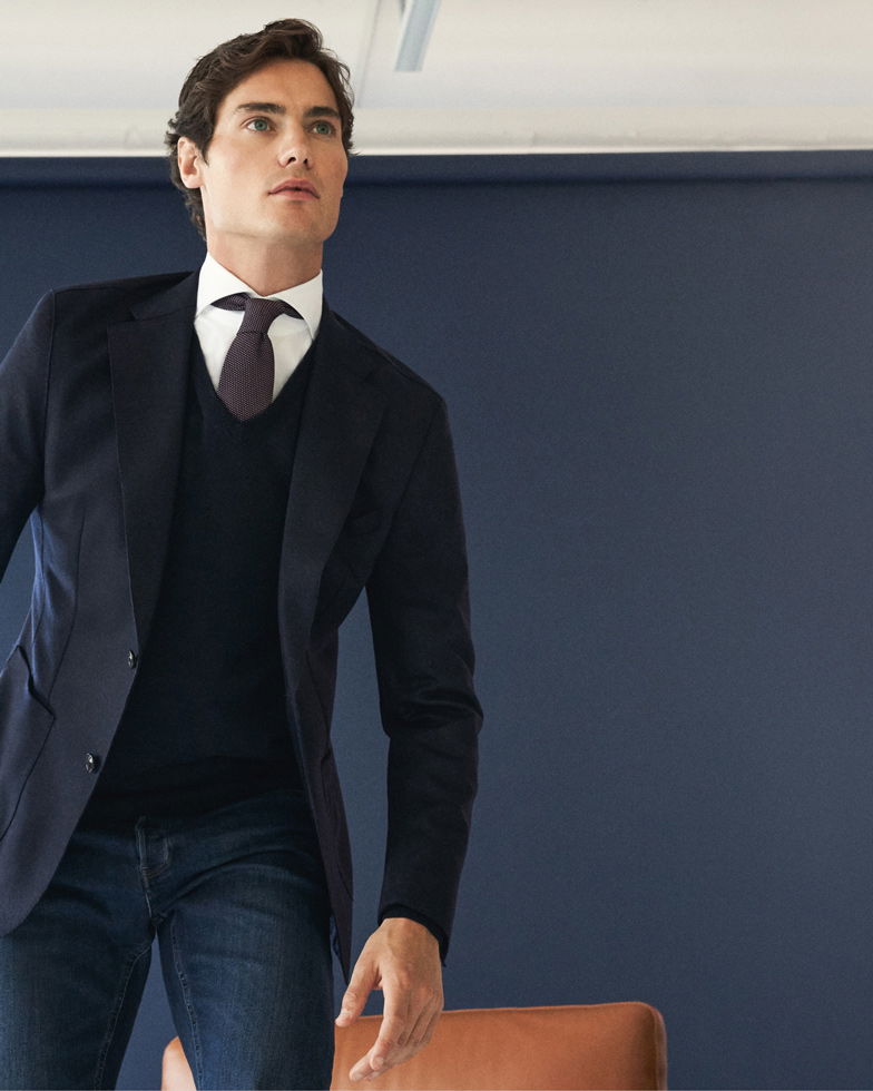 Mann trägt navy Sakko, blauen V-Neck Strickpullover, weißes Hemd, dunkle Krawatte und Jeans vor blauem Hintergrund.