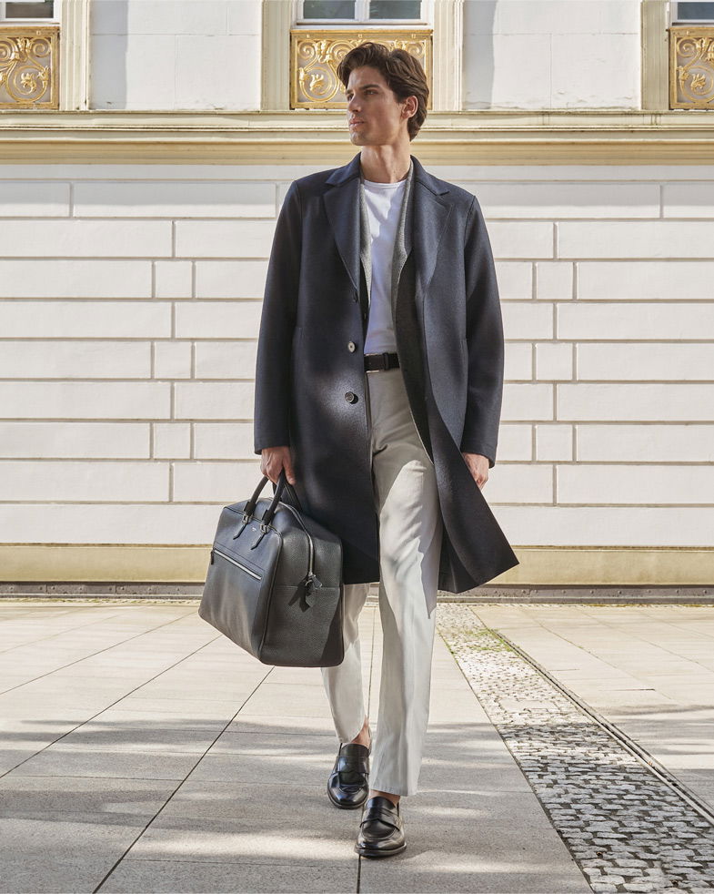 Mann trägt schwarzen Wollmantel, weißes T-Shirt, beige Hose, schwarze Loafer und hält schwarze Weekender-Tasche in der Hand.