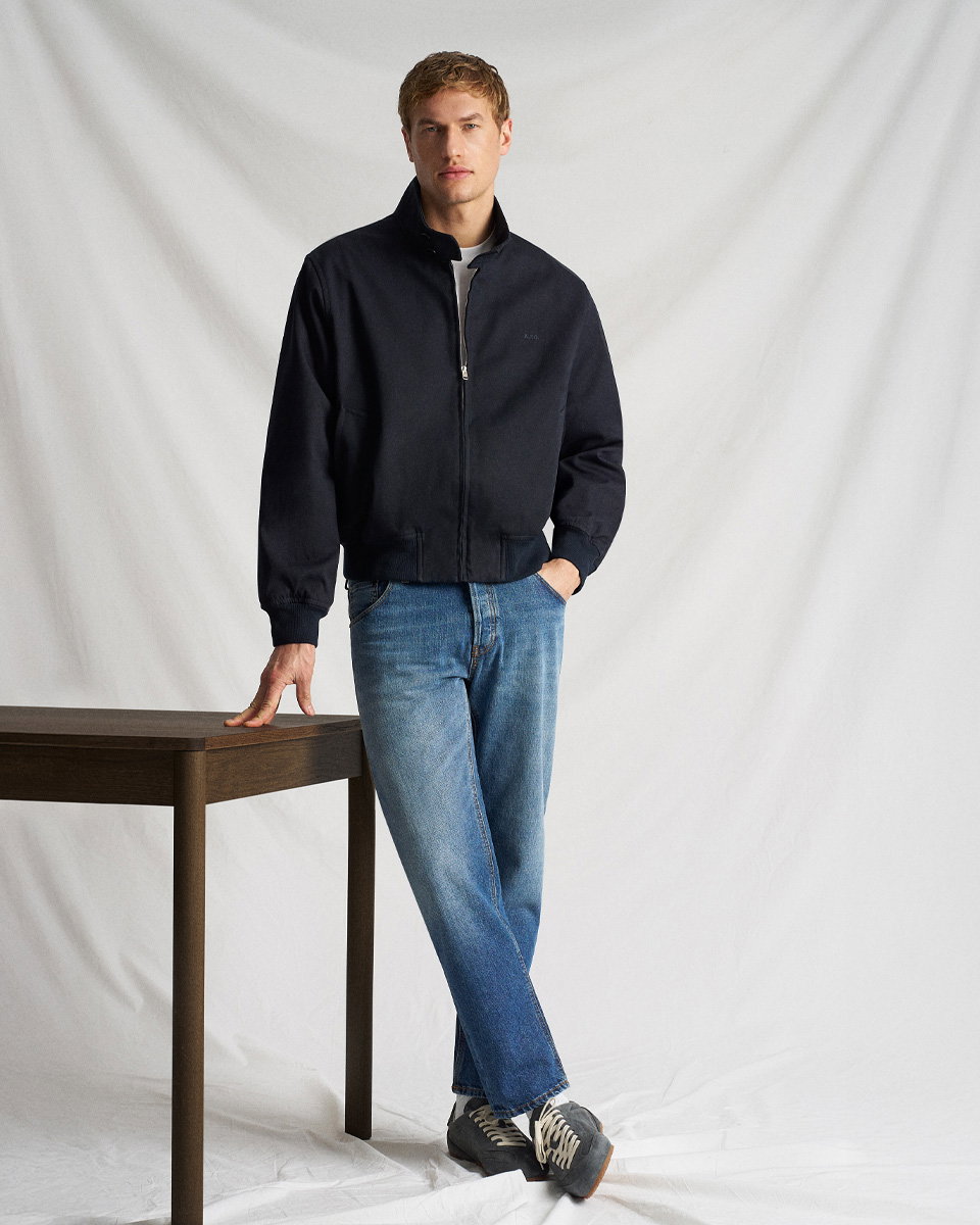 Mann mit blauer Jeans, grauem Shirt, navy Blouson und blauen Sneakern steht mit einer Hand an einem Holztisch vor weißem Hintergrund.