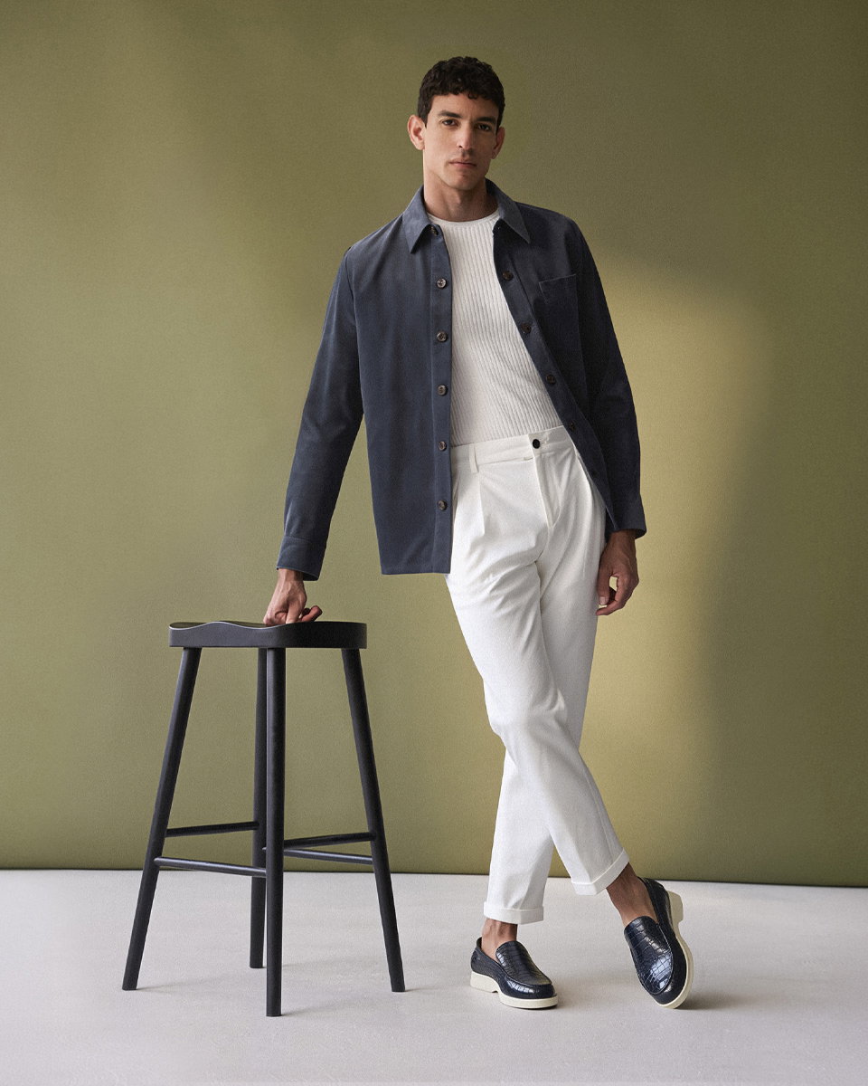 Mann lehnt an schwarzem Hocker, trägt offwhite Hose, weißes Shirt, dunkelblaues Overshirt und navy Loafer, grüner Hintergrund.