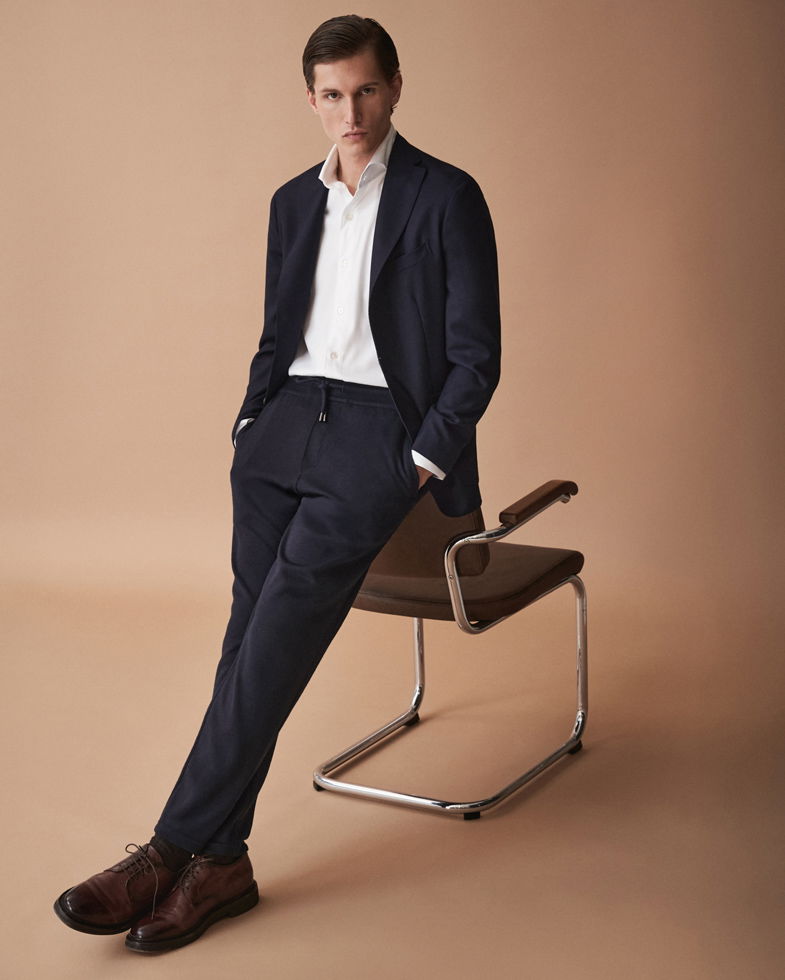 Mann lehnt an Stuhl, trägt navy Sakko, weiße Hemd, Jogginghose und braune Schnürschuhe vor beigem Hintergrund.