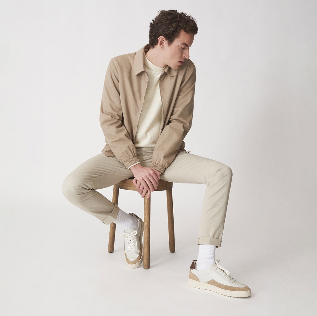 Mann sitzt auf Holz-Hocker, trägt beige Hose, beige Jacke, cremefarbigen Pullover und weiße Sneaker mit beigen Akzenten. Mann sitzt auf Holz-Hocker, trägt beige Hose, beige Jacke, cremefarbigen Pullover und weiße Sneaker mit beigen Akzenten.
