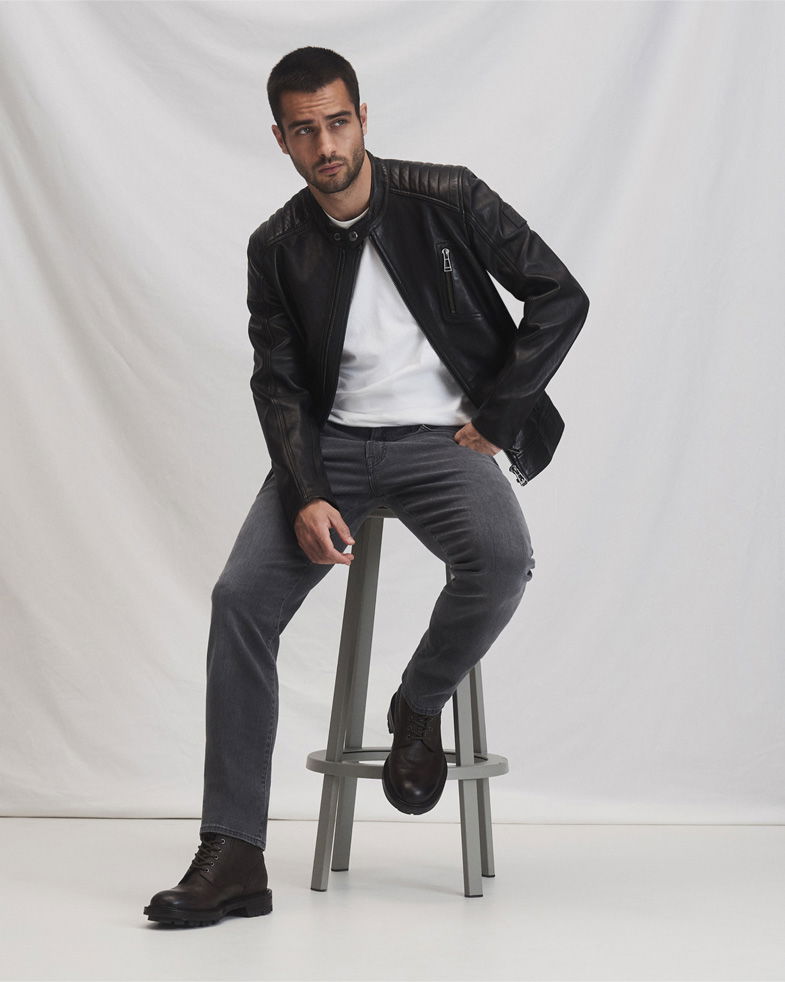 Mann sitzt auf grauem Hocker, trägt schwarze Lederjacke, weißes T-Shirt, graue Jeans und schwarze Schnürboots.