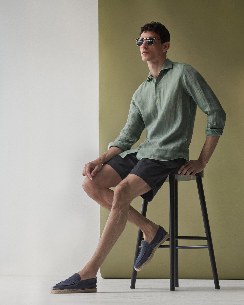 Mann sitzt auf schwarzem Hocker, trägt salbeigrünes Hemd, navy blaue Shorts, blaue Espadrilles und Sonnenbrille.