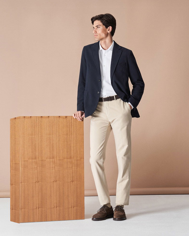 Mann lehnt mit linker Hand an Holzkiste, trägt navy Sakko, weißes Hemd, beige Hose und braune Schuhe, schaut seitlich.