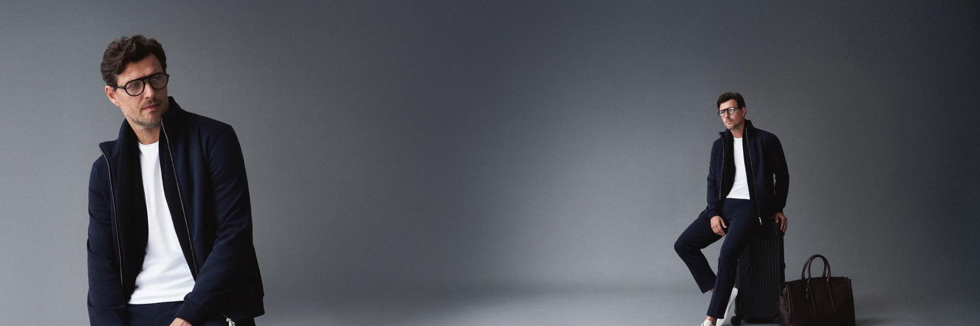 Mann mit dunkelblauer Jogginghose, Sweatjacke, weißem Shirt und weißen Sneakern sitzt auf schwarzem Koffer, braune Tasche daneben. Mann mit dunkelblauer Jogginghose, Sweatjacke, weißem Shirt und weißen Sneakern sitzt auf schwarzem Koffer, braune Tasche daneben.