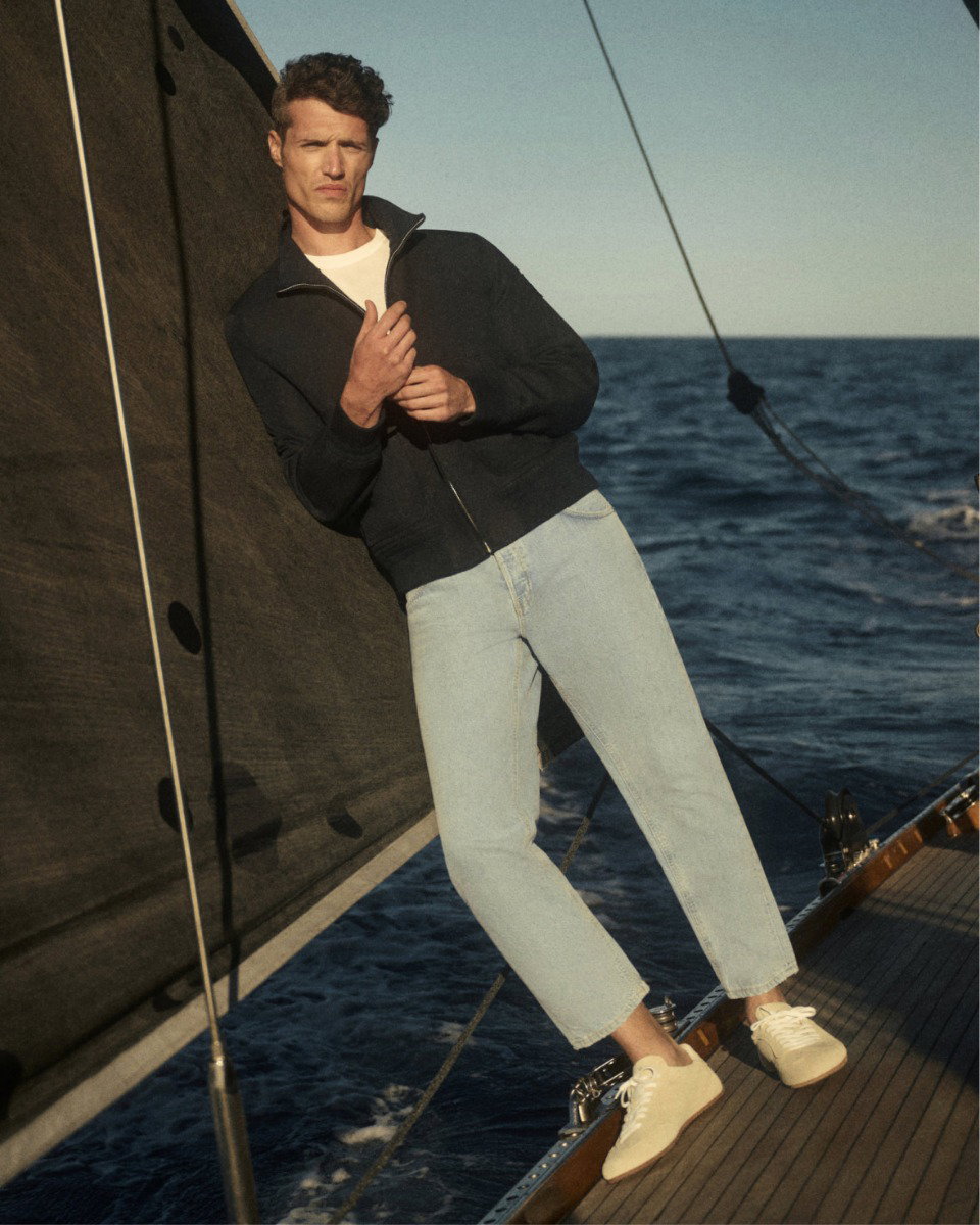 Mann steht auf Segelboot, trägt weiße Sneaker, helle Jeans, weißes Shirt und navy Blouson, Meer und Segel im Hintergrund.