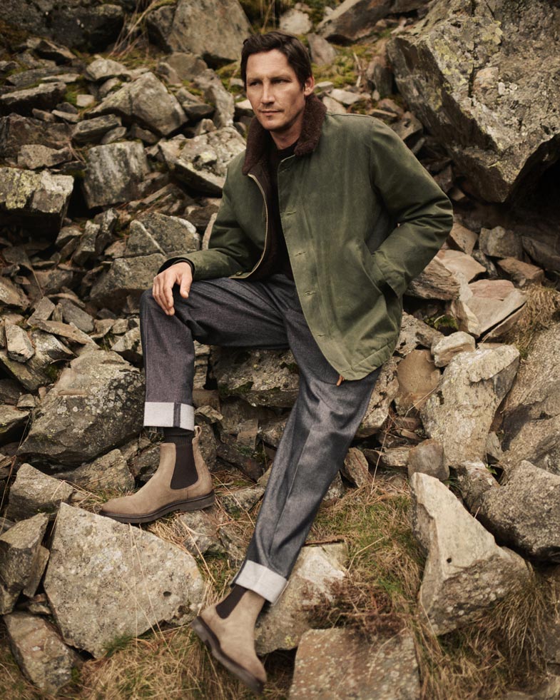 Mann sitzt auf Steinen, trägt olivgrüne Jacke mit Fellkragen, dunkelgraue Hose, beige Boots und schwarzes Shirt.