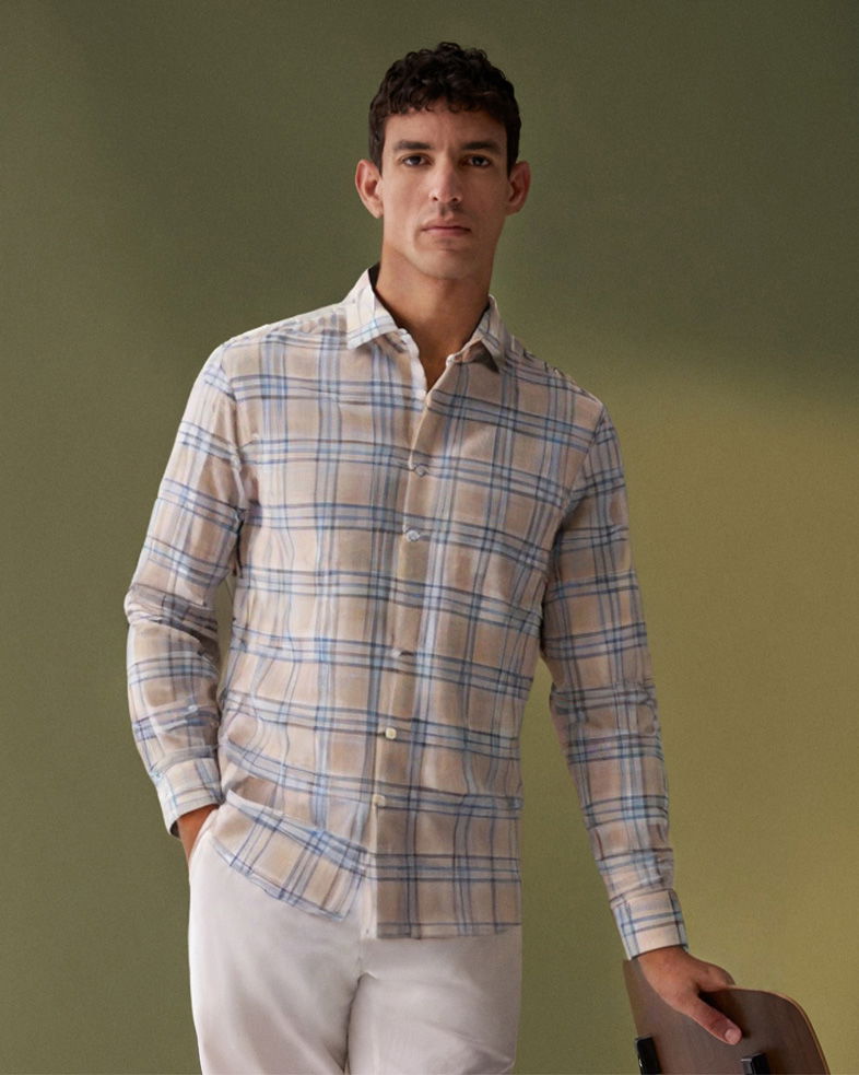 Herrenportrait, stehend, mit beige-blau kariertem Hemd und weißer Hose, eine Hand in der Hosentasche, vor grünem Hintergrund.