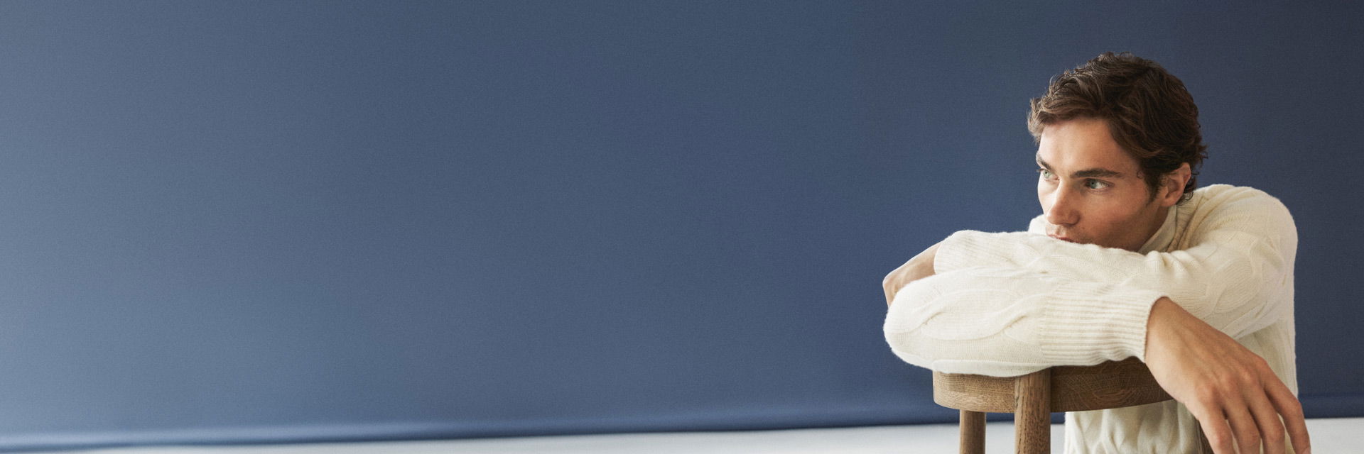 Mann sitzt rückwärts auf Holzstuhl, stützt Arme auf Lehne, trägt cremefarbenen Rollkragenpullover, blickt nach rechts. Mann sitzt rückwärts auf Holzstuhl, stützt Arme auf Lehne, trägt cremefarbenen Rollkragenpullover, blickt nach rechts.