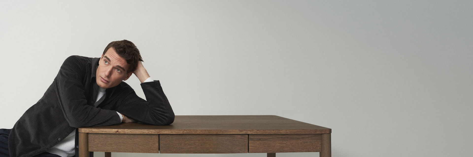 Mann lehnt mit schwarzem Cord-Overshirt und weißem Pullover entspannt mit Arm auf braunem Holztisch vor hellgrauer Wand. Mann lehnt mit schwarzem Cord-Overshirt und weißem Pullover entspannt mit Arm auf braunem Holztisch vor hellgrauer Wand.