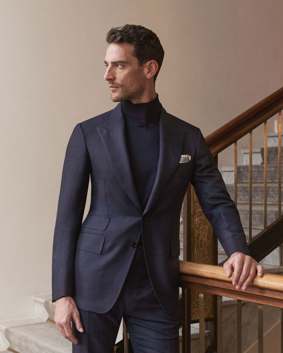 Herrenportrait auf Treppe, navy gemusterter Anzug, navy Rollkragenpullover, weißes Einstecktuch, Hand am Geländer.