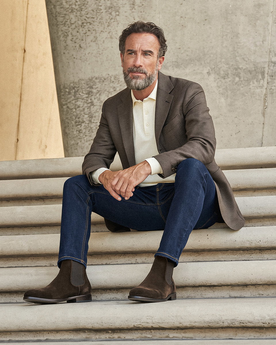 Mann mit grauem Bart sitzt auf Treppe, trägt braunes Sakko, cremefarbenes Langarmpolo, blaue Jeans und braune Boots.