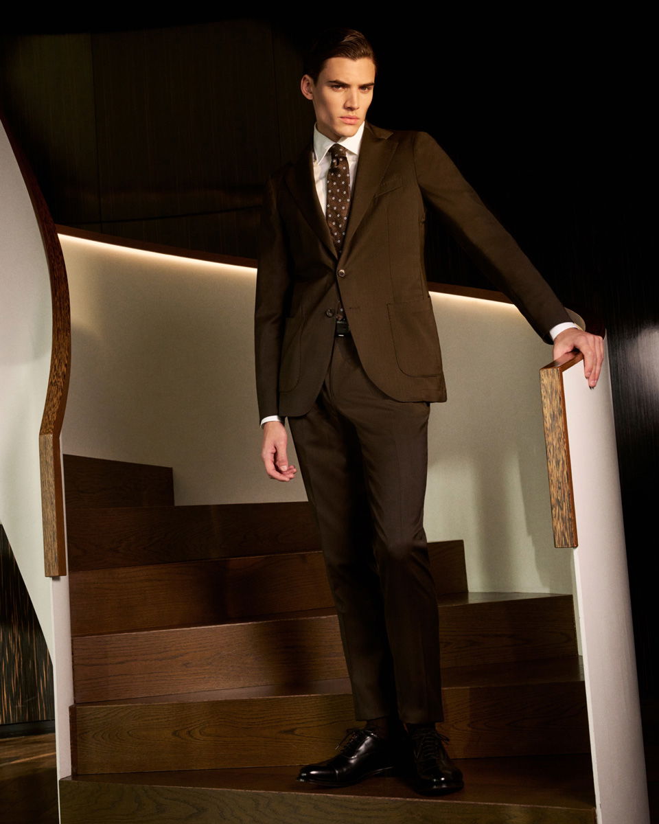 Mann steht auf einer Treppe, trägt braunen Anzug, weißes Hemd und braun gemusterte Krawatte, lehnt an Holzgeländer. Mann steht auf einer Treppe, trägt braunen Anzug, weißes Hemd und braun gemusterte Krawatte, lehnt an Holzgeländer.