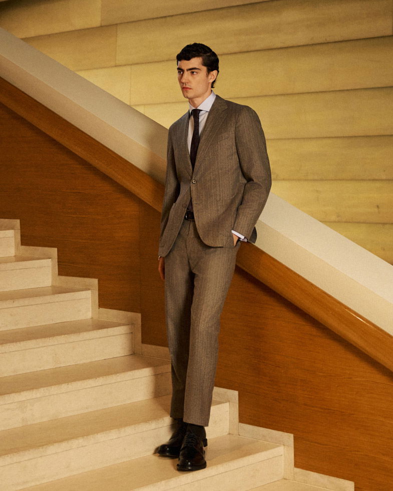 Mann im grauen Anzug, weißem Hemd und schwarzer Krawatte steht auf heller Treppe neben Holzgeländer in Innenraum. Mann im grauen Anzug, weißem Hemd und schwarzer Krawatte steht auf heller Treppe neben Holzgeländer in Innenraum.