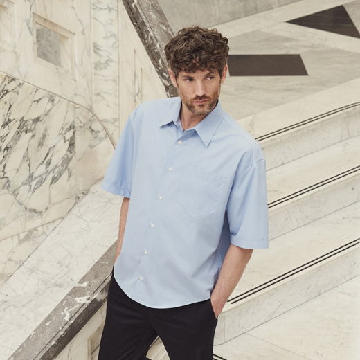 Mann mit lockigem Haar trägt hellblaues Kurzarmhemd und schwarze Hose, steht auf weißer Steintreppe mit Marmorgeländer.