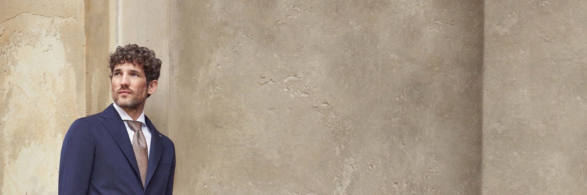 Mann mit lockigen Haaren trägt dunkelblauen Anzug, weißes Hemd und gemusterte braune Krawatte, lehnt an hellem Betonpfeiler. Mann mit lockigen Haaren trägt dunkelblauen Anzug, weißes Hemd und gemusterte braune Krawatte, lehnt an hellem Betonpfeiler.