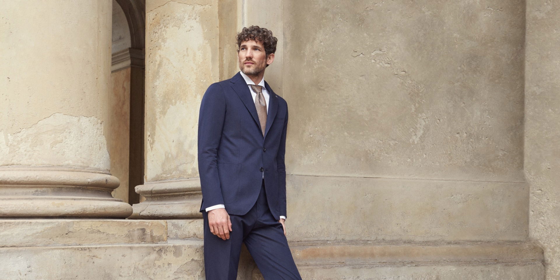 Mann mit lockigem Haar trägt dunkelblauen Anzug, weißes Hemd und braune gemusterte Krawatte lehnt an historischem Säulengang.