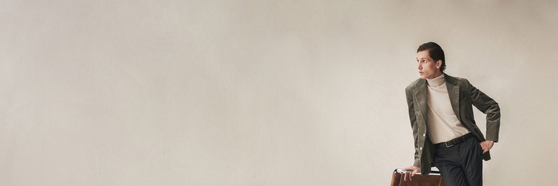 Mann lehnt an Stuhl, trägt cremefarbenen Rollkragenpullover, olivfarbenes Cordsakko und dunkle Hose vor beigem Hintergrund. Mann lehnt an Stuhl, trägt cremefarbenen Rollkragenpullover, olivfarbenes Cordsakko und dunkle Hose vor beigem Hintergrund.