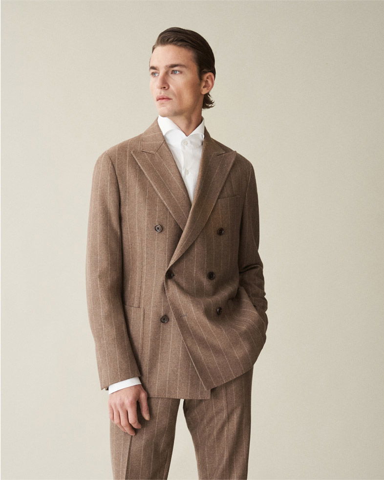 Mann mit braunem, gestreiftem Anzug und weißem Hemd steht vor beigem Hintergrund, eine Hand in der Hosentasche.