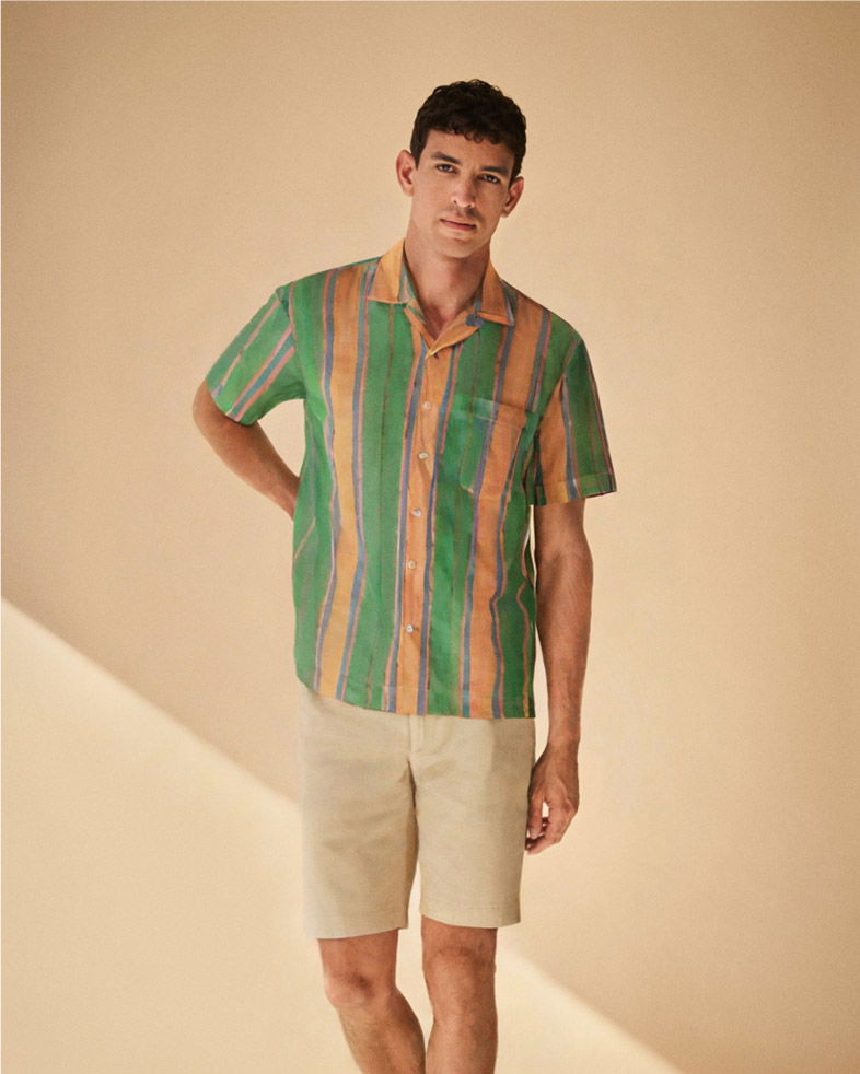 Mann mit kurzem, gestreiftem Hemd in Orange, Grün, Blau und beige Shorts vor hellem, einfarbigem Hintergrund.