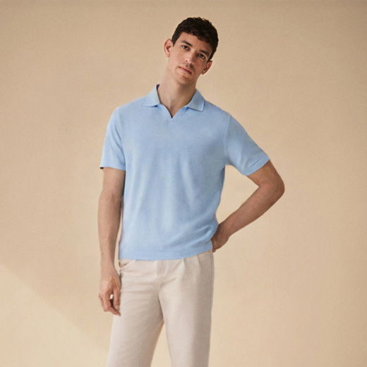 Mann mit kurzem dunklem Haar trägt hellblaues Strickpolo und beige Hose vor sandfarbenem Hintergrund, steht leicht seitlich.