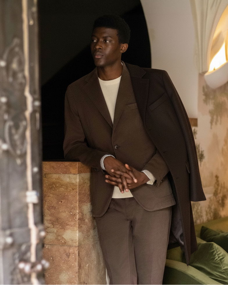 Mann lehnt an Mauer, trägt braunen Anzug, cremefarbenen Rundhalspullover, dunklen Mantel über der Schulter, nach links schauend. Mann lehnt an Mauer, trägt braunen Anzug, cremefarbenen Rundhalspullover, dunklen Mantel über der Schulter, nach links schauend.