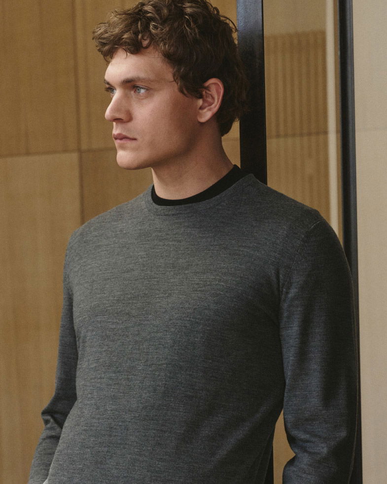 Mann mit kurzen lockigen Haaren trägt grauen Feinstrickpullover aus Merinowolle, steht seitlich vor Holz- und Glaswand. Mann mit kurzen lockigen Haaren trägt grauen Feinstrickpullover aus Merinowolle, steht seitlich vor Holz- und Glaswand.