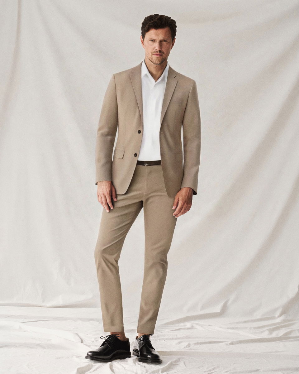 Mann im beigen Anzug mit weißem Hemd und schwarzen Schnürschuhen steht vor hellem Hintergrund im Fotostudio.