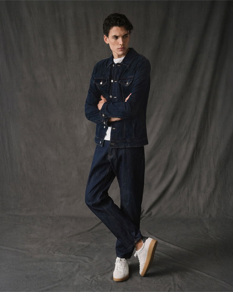 Mann im dunkelblauen Denim-Jacke und Hose, weißem T-Shirt und weißen Sneakern vor grauem Hintergrund stehend.