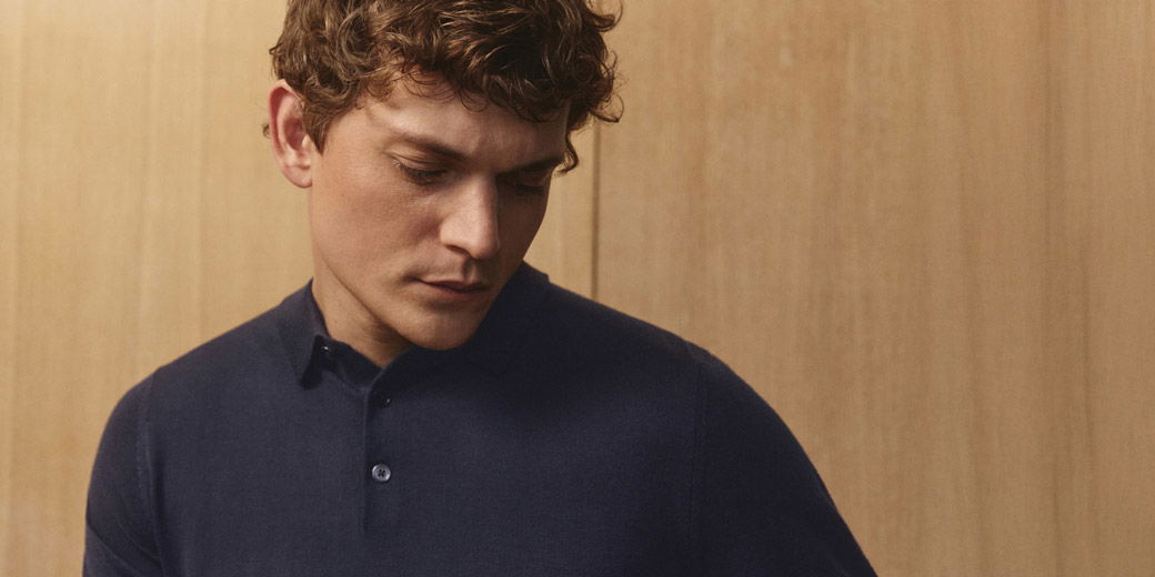 Junger Mann mit kurzen lockigen Haaren trägt dunkelblaues Merino-Strickpolo vor hellem Holzpaneel. Junger Mann mit kurzen lockigen Haaren trägt dunkelblaues Merino-Strickpolo vor hellem Holzpaneel.