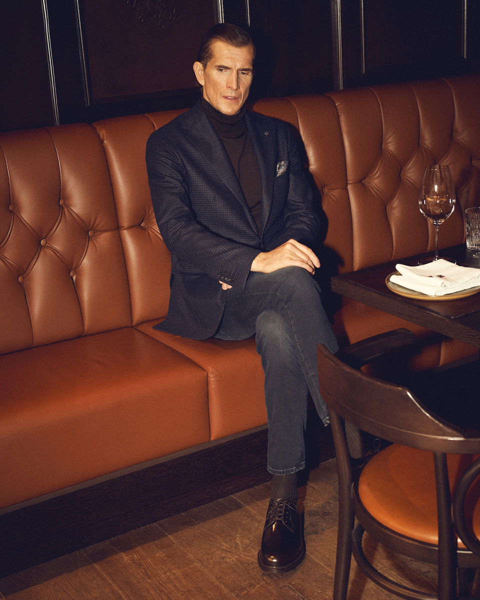 Mann mit blauem Sakko, schwarzem Rollkragenpullover, grauer Jeans und braunen Schnürschuhen sitzt in brauner Ledercouch im Restaurant. Mann mit blauem Sakko, schwarzem Rollkragenpullover, grauer Jeans und braunen Schnürschuhen sitzt in brauner Ledercouch im Restaurant.