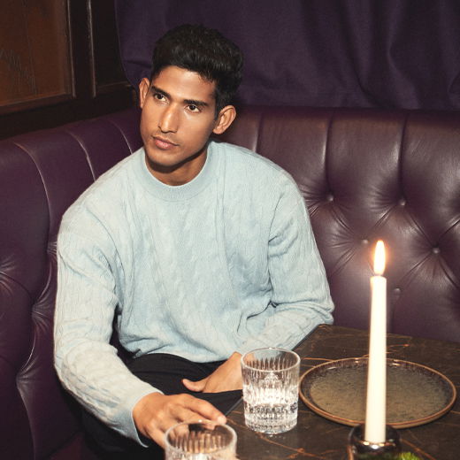 Mann mit dunkelbraunen Haaren sitzt in lila Lederbank, trägt hellblauen Strickpullover, vor ihm Kerze, Glas und Teller auf Tisch. Mann mit dunkelbraunen Haaren sitzt in lila Lederbank, trägt hellblauen Strickpullover, vor ihm Kerze, Glas und Teller auf Tisch.