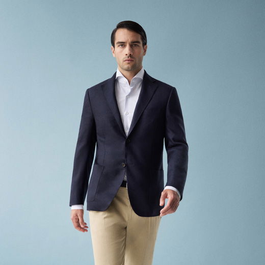 Mann trägt navy Sakko, weißes Hemd und beige Hose vor hellblauem Hintergrund, schaut ernst und geht leicht nach vorn.