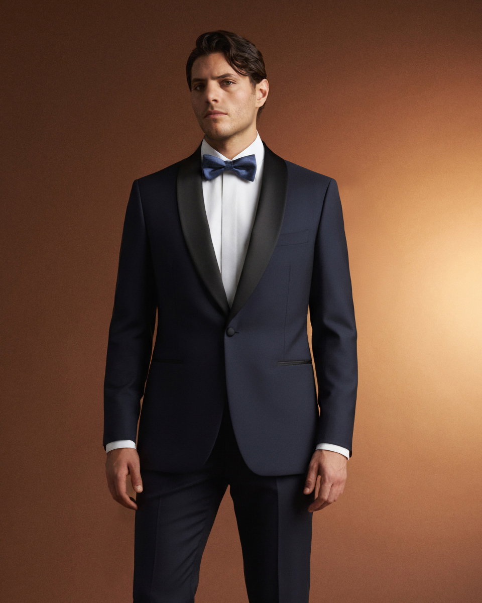 Herr in navyblauem Smoking mit schwarzem Revers, weißem Hemd und blauer Fliege vor rostbraunem Hintergrund.