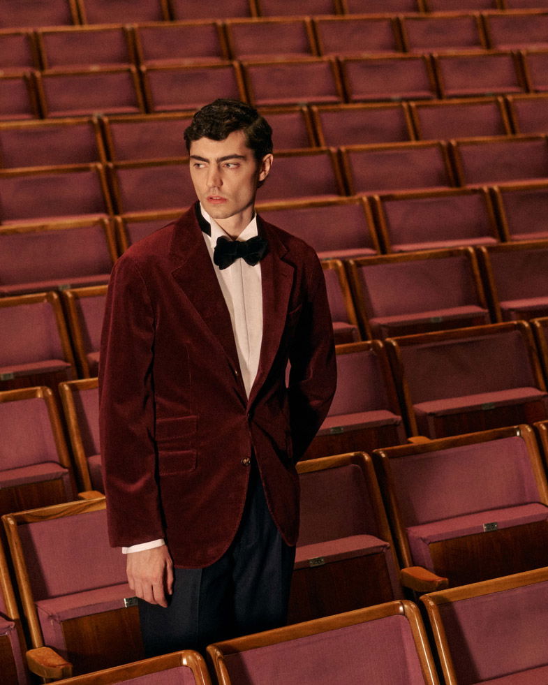 Mann im roten Samtsakko, weißem Hemd und schwarzer Fliege steht in einem leeren Theatersaal mit roten Sitzreihen. Mann im roten Samtsakko, weißem Hemd und schwarzer Fliege steht in einem leeren Theatersaal mit roten Sitzreihen.