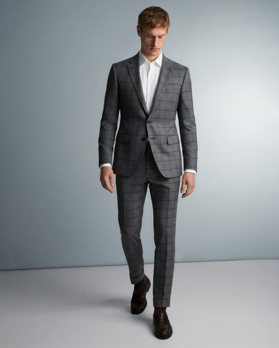 Mann trägt grauen karierten Anzug, weißes Hemd und braune Schuhe, schaut nach unten, steht vor hellgrauem Hintergrund.