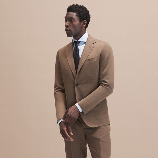 Mann trägt hellbraunen Anzug, pastellblaues Hemd und braun gemusterte Krawatte, vor beige Hintergrund stehend.