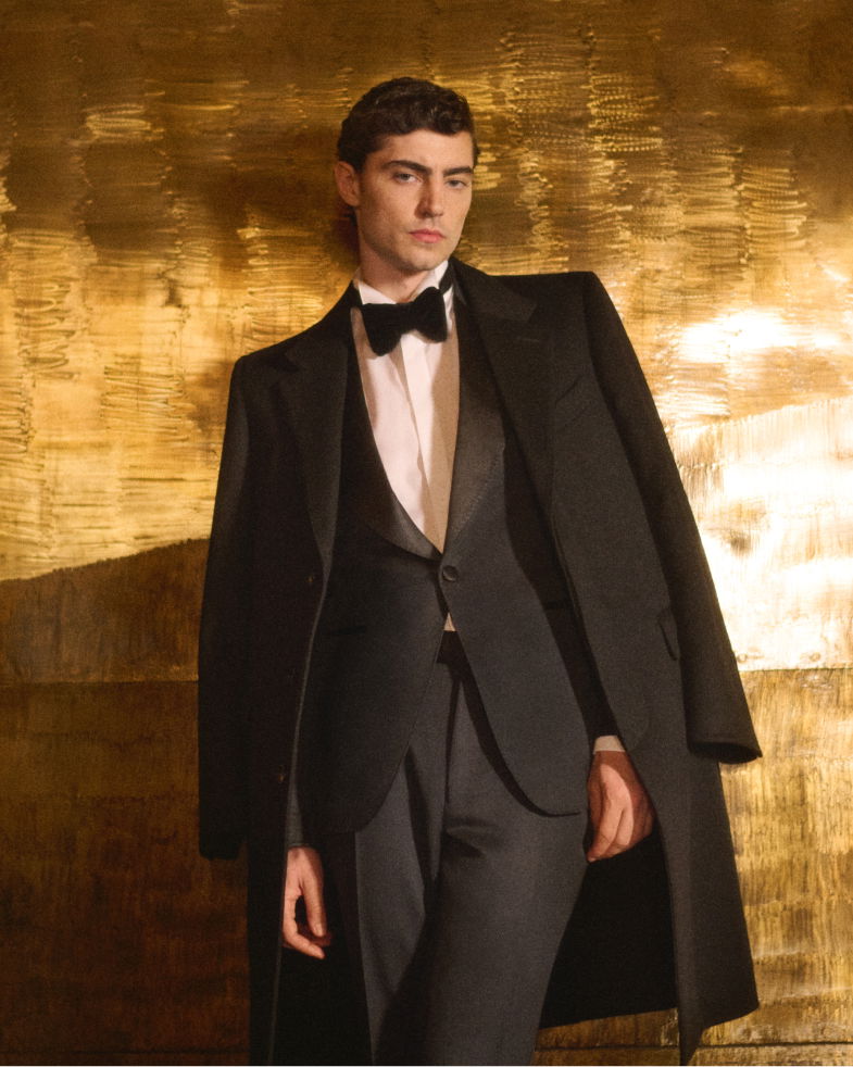 Mann trägt schwarzen Smoking mit weißem Hemd, schwarzer Fliege und schwarzem Mantel vor goldener Wand. Mann trägt schwarzen Smoking mit weißem Hemd, schwarzer Fliege und schwarzem Mantel vor goldener Wand.