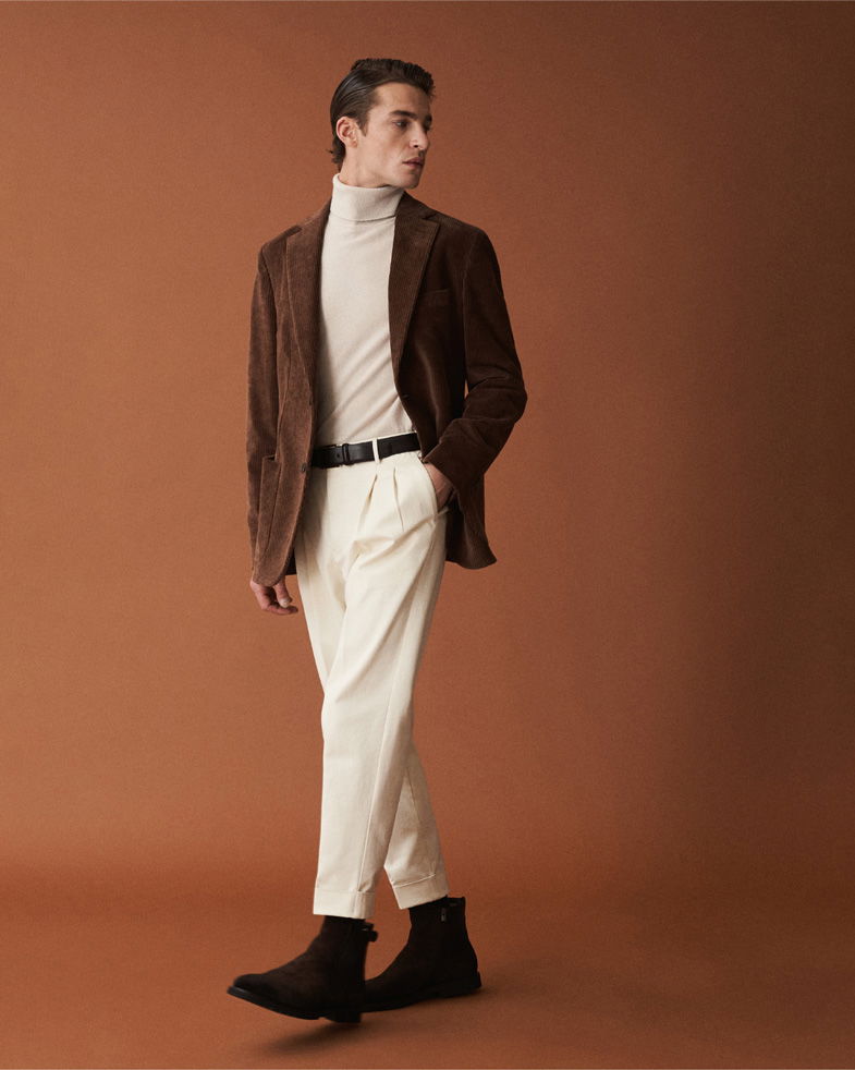 Herr in braunem Cordsakko, beigem Rollkragenpullover, cremefarbener Hose und braunen Boots vor braunem Hintergrund.