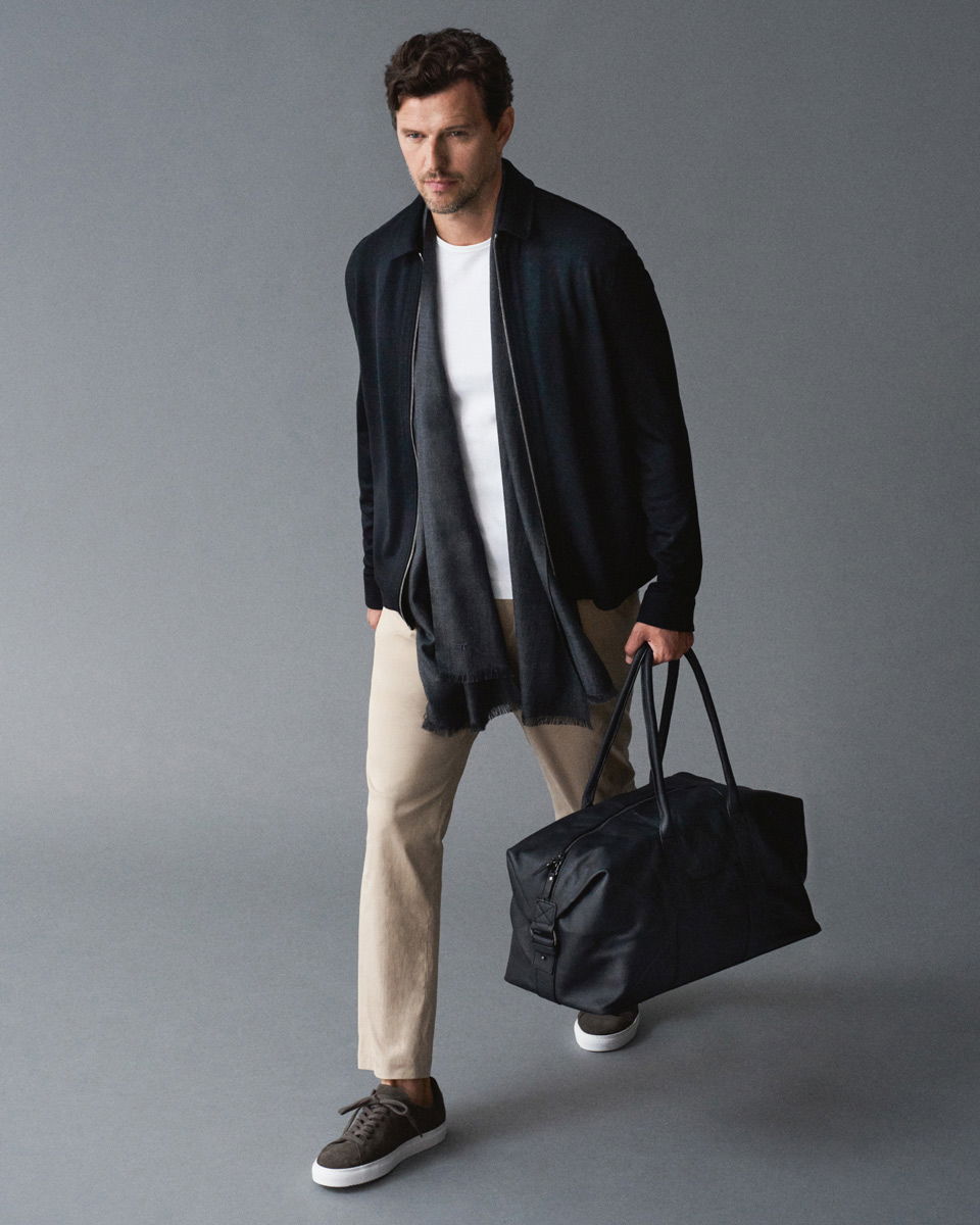 Mann in navy Jacke, weißem Shirt, beiger Hose, schwarzem Schal und Sneakern geht mit schwarzem Weekender in grauem Raum.