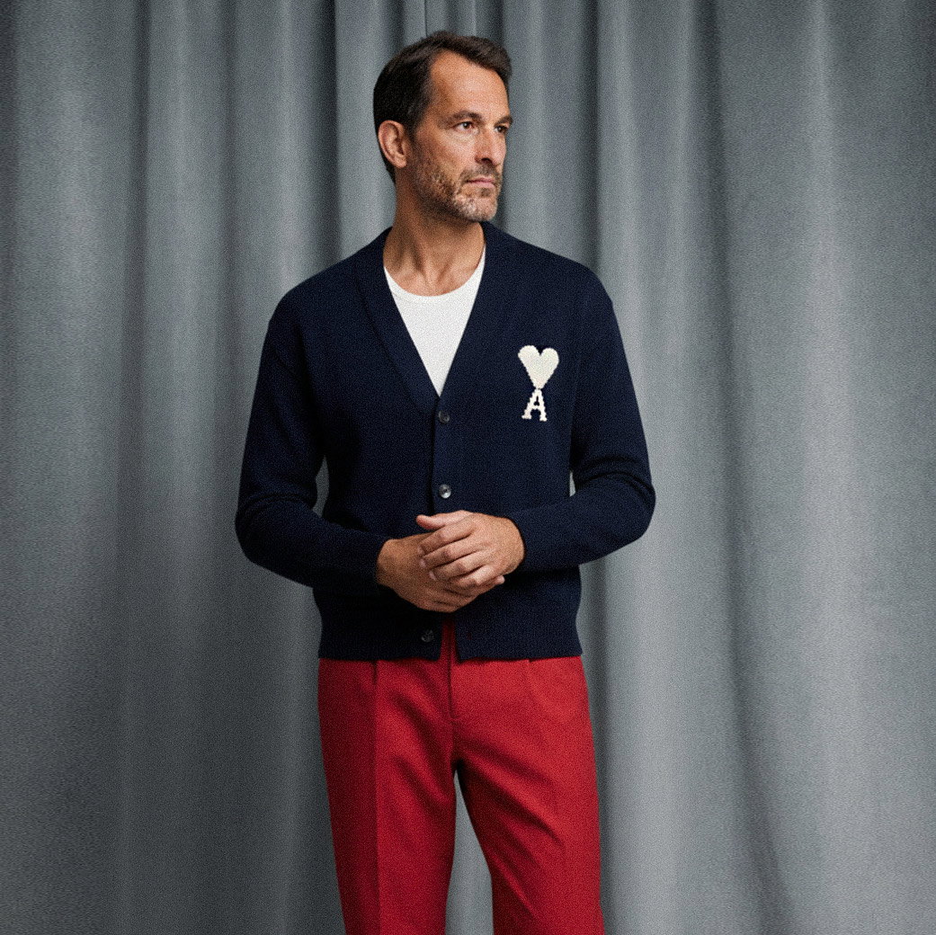 Mann vor graublauem Hintergrund trägt navy Cardigan mit weißem Herz und A, weißes Rundhalsshirt und rote Chinohose. Mann vor graublauem Hintergrund trägt navy Cardigan mit weißem Herz und A, weißes Rundhalsshirt und rote Chinohose.