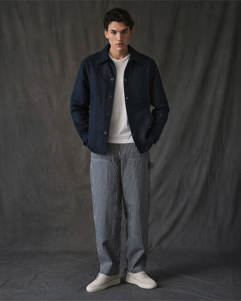 Mann trägt weiße Sneakers, blau-weiß gestreifte Loose-Fit-Hose, weißes T-Shirt und dunkelblaues Overshirt vor grauem Hintergrund.