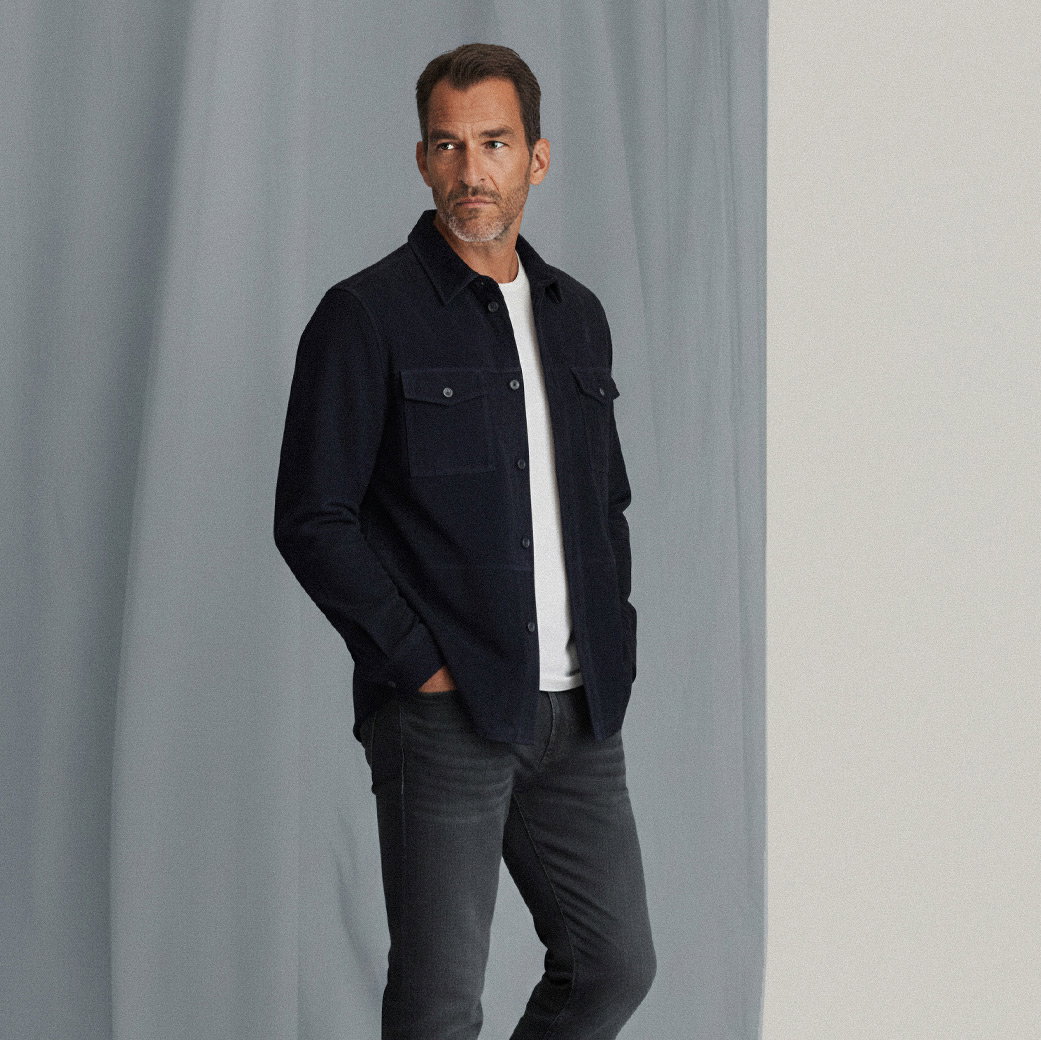 Mann steht vor grauem Hintergrund, trägt navy Overshirt, weißes T-Shirt und dunkle Jeans, Hände in den Taschen. Mann steht vor grauem Hintergrund, trägt navy Overshirt, weißes T-Shirt und dunkle Jeans, Hände in den Taschen.