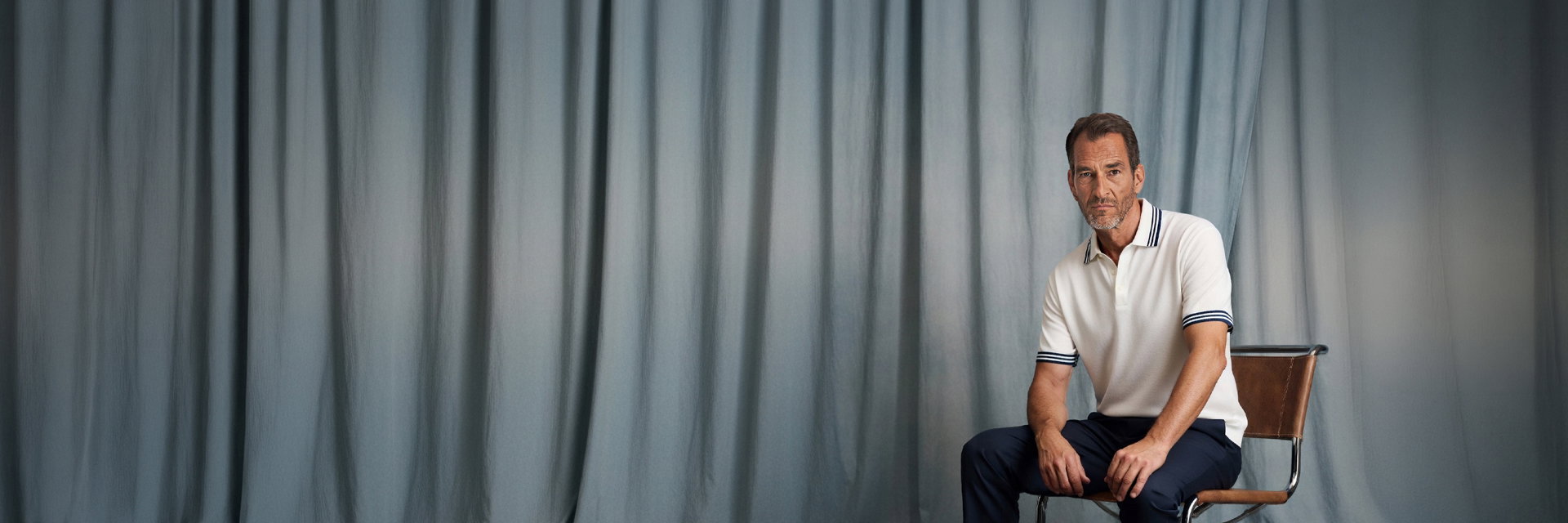 Mann im weißen Poloshirt mit dunkelblauen Streifen sitzt auf braunem Stuhl vor graublauem Vorhang, ernst blickend. Mann im weißen Poloshirt mit dunkelblauen Streifen sitzt auf braunem Stuhl vor graublauem Vorhang, ernst blickend.