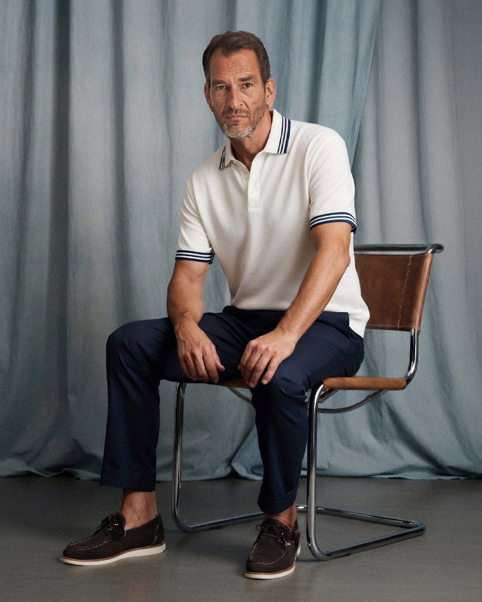 Mann sitzt auf Stuhl vor blauem Vorhang, trägt weißes Poloshirt mit Streifen, dunkelblaue Hose und braune Bootsschuhe. Mann sitzt auf Stuhl vor blauem Vorhang, trägt weißes Poloshirt mit Streifen, dunkelblaue Hose und braune Bootsschuhe.