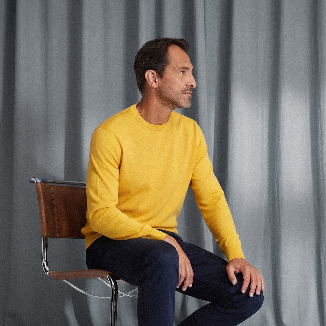 Mann sitzt auf Stuhl vor graublauem Vorhang, trägt gelben Rundhalspullover und dunkelblaue Hose, schaut nach rechts. Mann sitzt auf Stuhl vor graublauem Vorhang, trägt gelben Rundhalspullover und dunkelblaue Hose, schaut nach rechts.