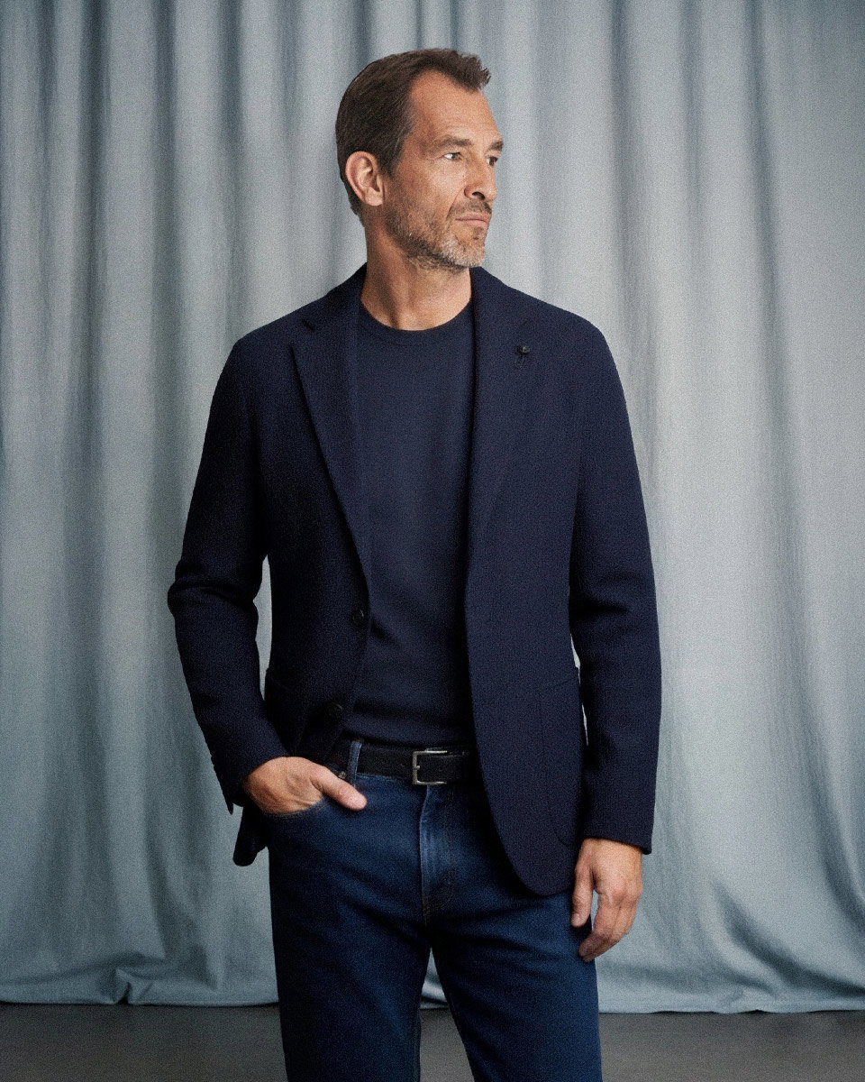 Mann mit braunem Haar und Bart trägt dunkelblaues Sakko, navy Rundhalspullover und dunkelblaue Jeans vor graublauem Hintergrund. Mann mit braunem Haar und Bart trägt dunkelblaues Sakko, navy Rundhalspullover und dunkelblaue Jeans vor graublauem Hintergrund.